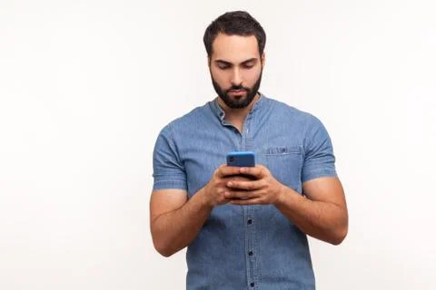 Assertive bearded man in blue shirt attentively looking at display of smartph Stock Photos