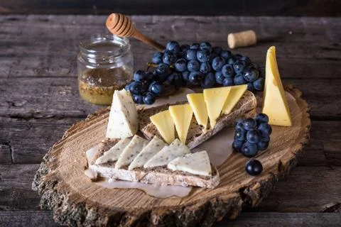 Assorted cheeses with grapes, bread, honey on dark wood background. Goat chee Stock Photos