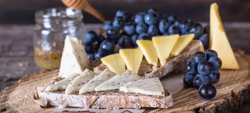 Assorted cheeses served with grapes, bread, honey, and dates on dark wood bac Stock Photos