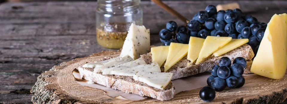 Assorted cheeses served with grapes, bread, honey, and dates on dark wood bac Stock Photos
