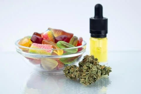 Assorted different gummy candies in a small glass bowl and a bottle of CBD ca Stock Photos