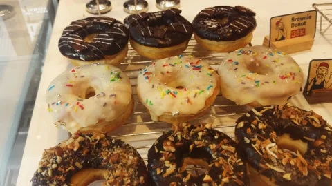 Assorted donuts on display in a bakery case, showcasing glazed, chocolate, brown Stock Footage 333115802