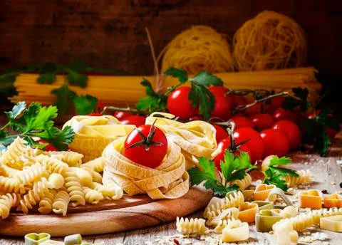Assorted dry pasta, cherry tomatoes and parsley on a vintage wooden backgroun Stock Photos