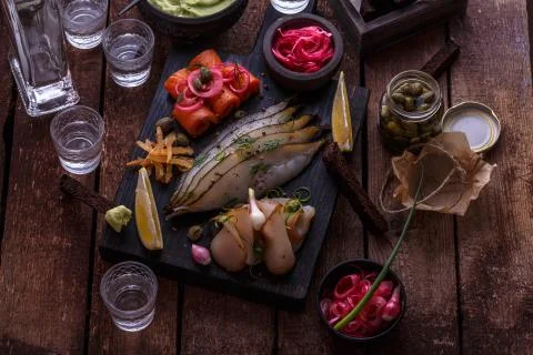 Assorted preserved fish on dark cutting board with vodka shots. Stock Photos