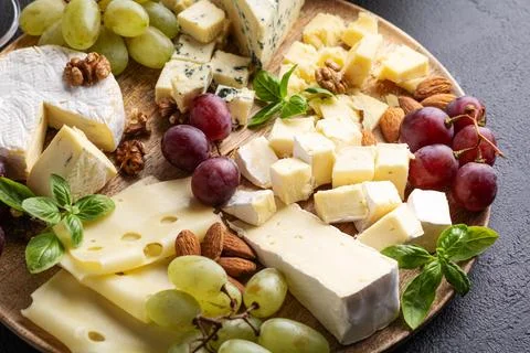 Assortment of Cheeses Grapes Nuts and Fresh Basil Leaves on Wooden Platter Stock Photos