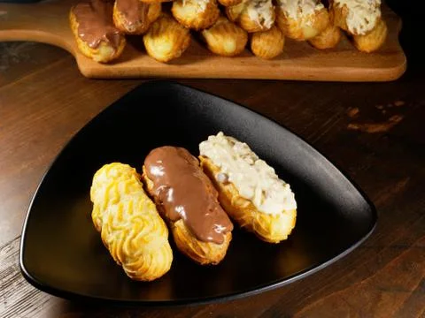 Assortment of eclairs on a plate. Different cakes on a black triangular plate. Stock Photos