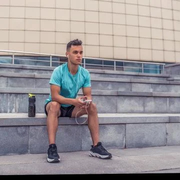 Athlete guy is resting, sitting on steps, listening music headphones, after Stock Photos