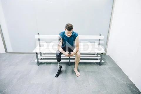 Athlete man with prosthetic leg sitting in gym locker room with mobile phone Stock Photos