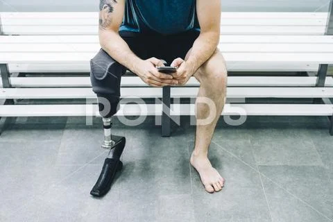 Athlete man with prosthetic leg sitting in gym locker room with mobile phone Stock Photos