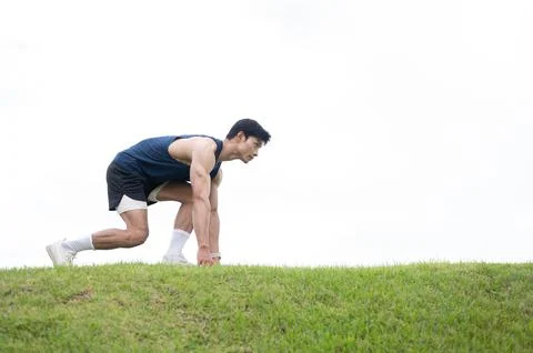 Athletic Asian man crouching preparing to run on the road over a grassy fie.. Stock Photos