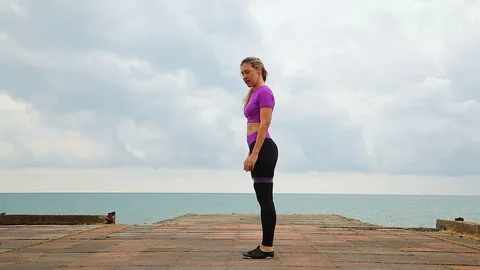 Athletic girl doing a warm up squat and jump with an elastic band near the sea 库存影片 114576643