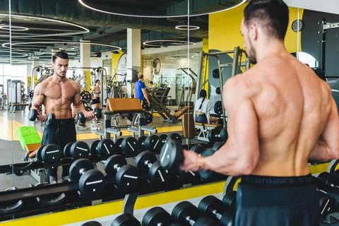 Athletic man doing dumbbell exercise in front of a mirror bare torso in the gym Stock Photos