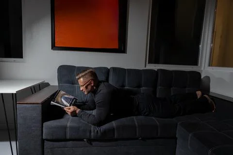Athletic man with glasses reading a book while lying on a dark sofa in a cozy Stock Photos