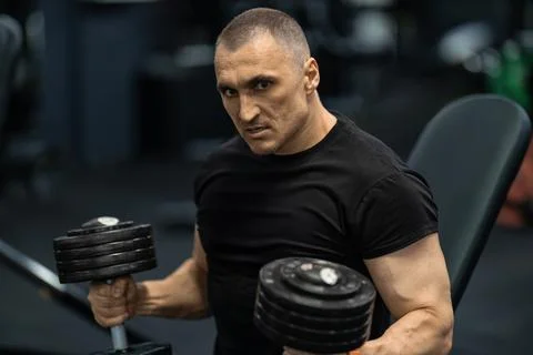 Athletic man lifting dumbbells while sitting on bench in gym focused on workout Stock Photos
