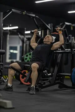 Athletic man performs bench press on shoulder press machine in gym during Stock Photos