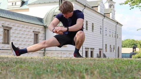 Athletic man stretches legs on grass in front of historic building during outdoo Stock Footage 315413830
