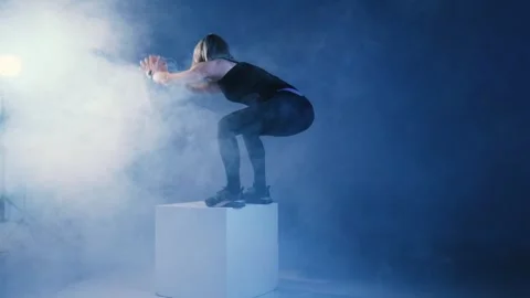 Athletic middle-aged woman doing sports exercise cube jump in smoky studio Stock Footage 268608965