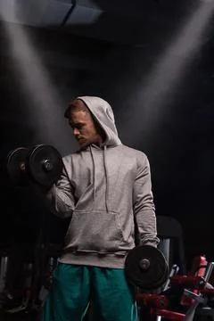 Athletic person performs dumbbell bicep curls in gym wearing gray hoodie during Stock Photos