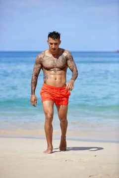 Athletic tattooed guy walking through the water at the tropical beach Stock Photos