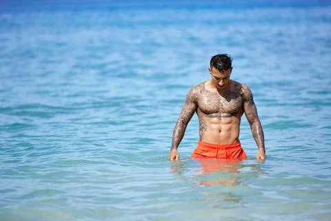 Athletic tattooed guy walking through the water at the tropical beach Stock Photos
