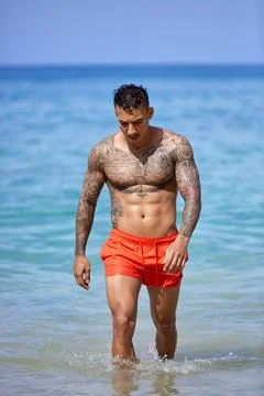 Athletic tattooed guy walking through the water at the tropical beach Stock Photos
