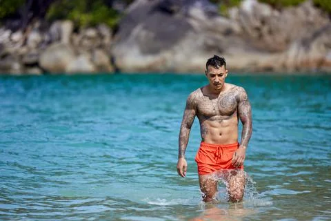 Athletic tattooed guy walking through the water at the tropical beach Stock Photos