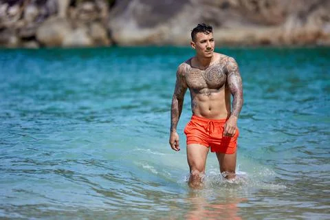 Athletic tattooed guy walking through the water at the tropical beach Stock Photos