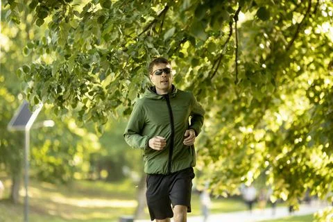 Athletic young man running while doing workout in sunny green park Stock Photos