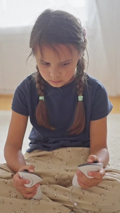 Attentive girl on lap comparing controllers, soft window light, braided pigtails Stock Footage 319966710