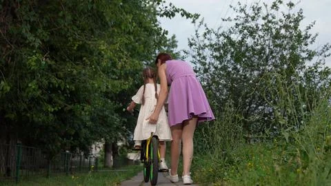 Attentive mother helps cute daughter to ride bicycle in city park backside view Stock Photos