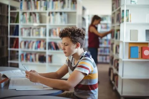 Attentive schoolboy using mobile phone while studying in library Stock Photos