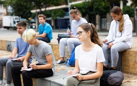 Attentive students write lesson in a notebook sitting on stone street parapet 库存照片