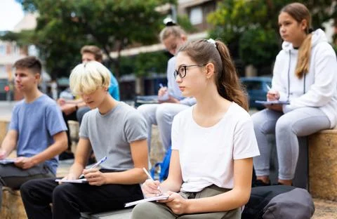 Attentive students write lesson in a notebook sitting on stone street parapet Foto stock