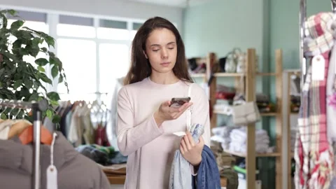 Attentive young girl scanning barcode on jeans pantaloons during shopping in Stock Footage 343715491