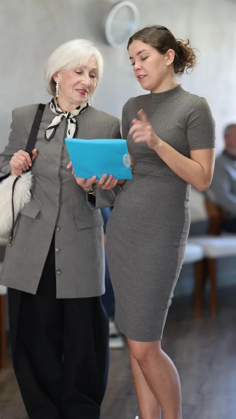 Attentive young woman explaining documents to retired woman while standing in Stock Footage 345584266