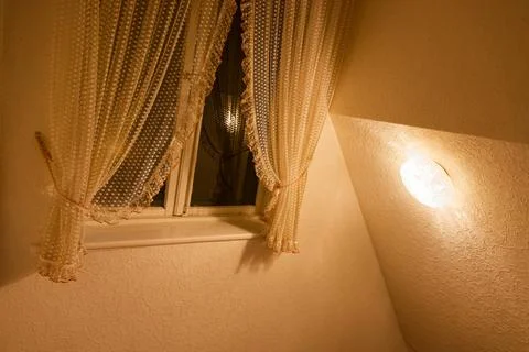 Attic window on slanted wall with ruffled curtains in soft light. Stock Photos
