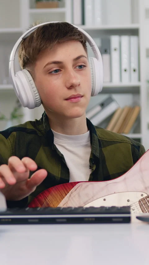 Attractive 13-14 years boy wearing headphones playing guitar looking at the lapt Stock Footage 333696266