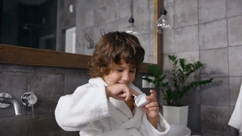 Attractive 5-aged boy with brown curly hair in robe playing with mom's cream in Stock Footage 120308378
