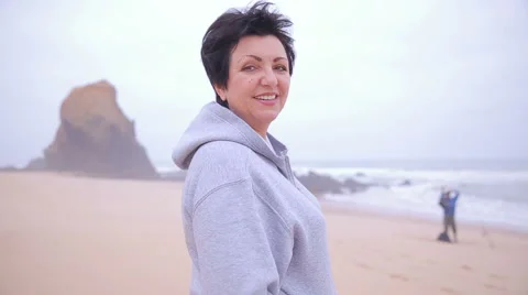 Attractive active mature woman standing on an ocean beach and smiling at camera. Stock Footage