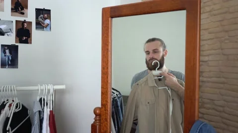 Attractive beard man is looking at himself into a mirror choosing a shirt Stock Footage 147893832