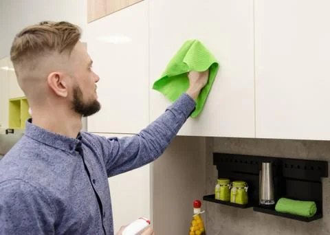 Attractive bearded man cleaning the handleless cabinet doors with a green rag Foto stock