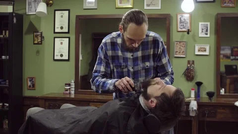 Attractive bearded man getting his beard shaved by modern barber in barber shop Stock Footage 69987441