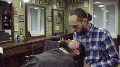 Attractive bearded man getting his beard shaved by modern barber in barber shop Stock Footage 69987667