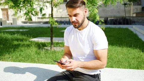 Attractive bearded man in white T-shirt sitting on the concrete wall and typing  Stock Footage 112363079