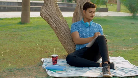 Attractive boy writing in a notebook while leaning against a tree in the park Stock Footage 154297270