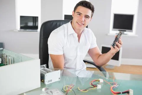 Attractive cheerful computer engineer sitting at desk holding hardware Stock Photos
