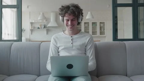 Attractive curly guy is sitting on couch in bright room home with laptop happy Stock Footage 158283812