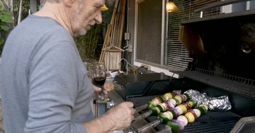 Attractive elderly senior man drinking red wine and grilling vegetable kebabs Stock Footage 86003076
