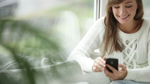 Attractive girl sitting by window using mobile phone looking at camera  Stock Footage 34150079