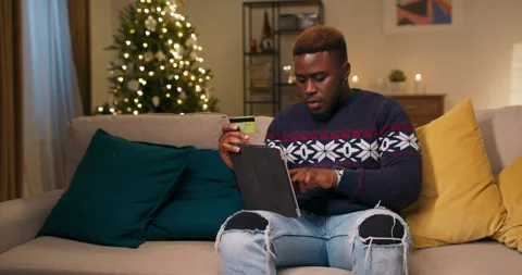 An attractive guy of African appearance sits on couch, holding tablet and bank 스톡 동영상 211100963